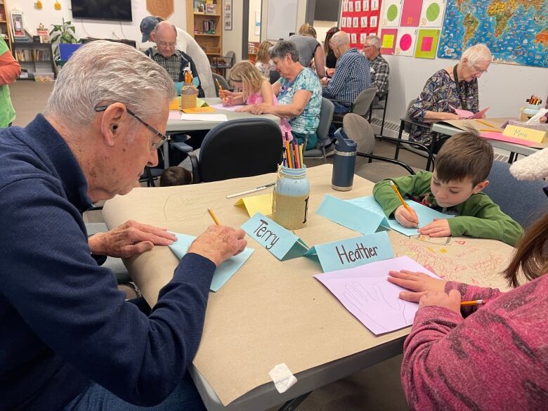 A senior man, on the left, draws on a piece of paper as he sits across a desk from young boy who also writes on a blue piece of paper.