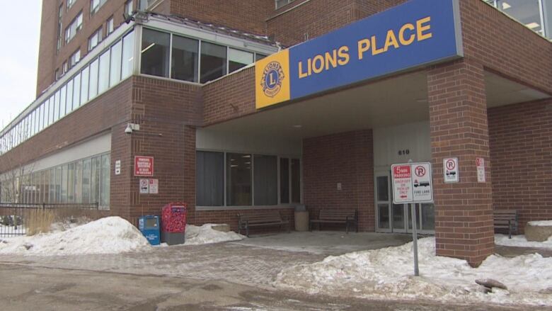 The bottom half of a tall brick apartment building is shown. It has a blue sign on it that says Lions Place. There is a post office back under a canopy entrance and several windows on the portion of the building to the left of the screen.