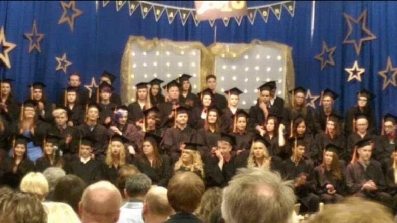 High school graduates are shown on a stage, in front of a blue curtain, with audience members in the foreground.