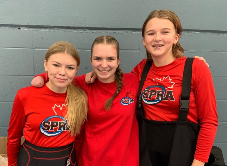 Three teenage girls wearing red shirts stand next to one another. They are smiling and the middle girls has her arms around the two girls next to her.