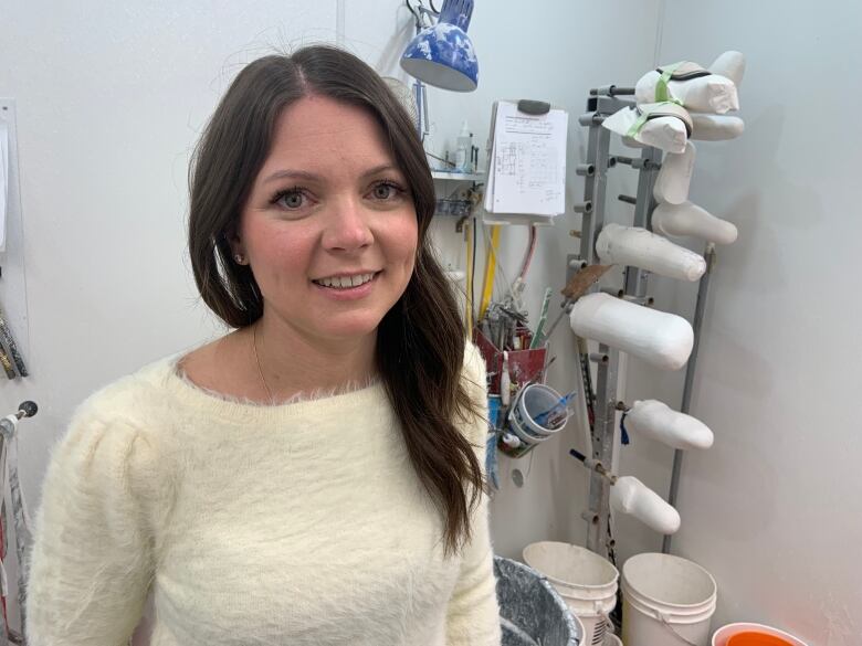 A woman with long brown hair, wearing a white sweater, stands in a white room. She is smiling. There are white plastic buckets and plaster legs behind her.