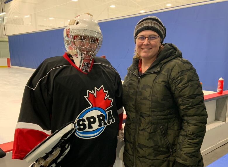 Two women stand side-by-side, in front of hockey ice, with their arms around one another. The young woman on the left is wearing full goalie gear, including a face mask. The woman on the right is wearing a green winter jacket.