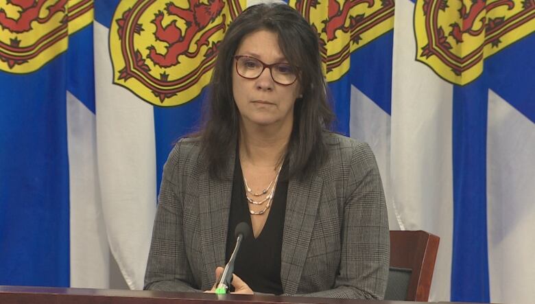 A woman with long dark hair and glasses sits at a podium.