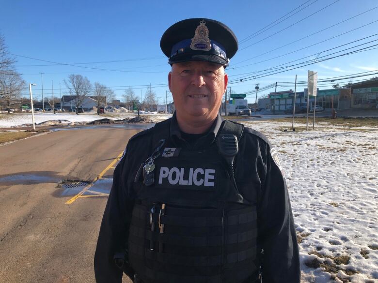 A man wearing a police uniform and hat stands in a parking lot smiling at the camera.