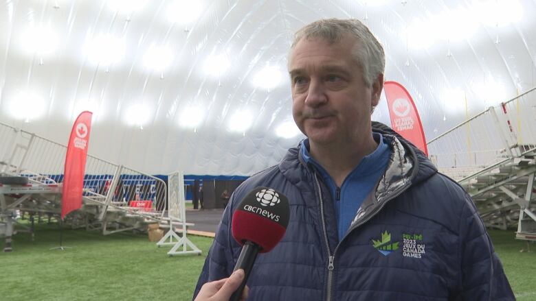 A man in a blue puffy jacket with a Canada Games 2023 logo speaks into a microphone, with bleachers in the background.