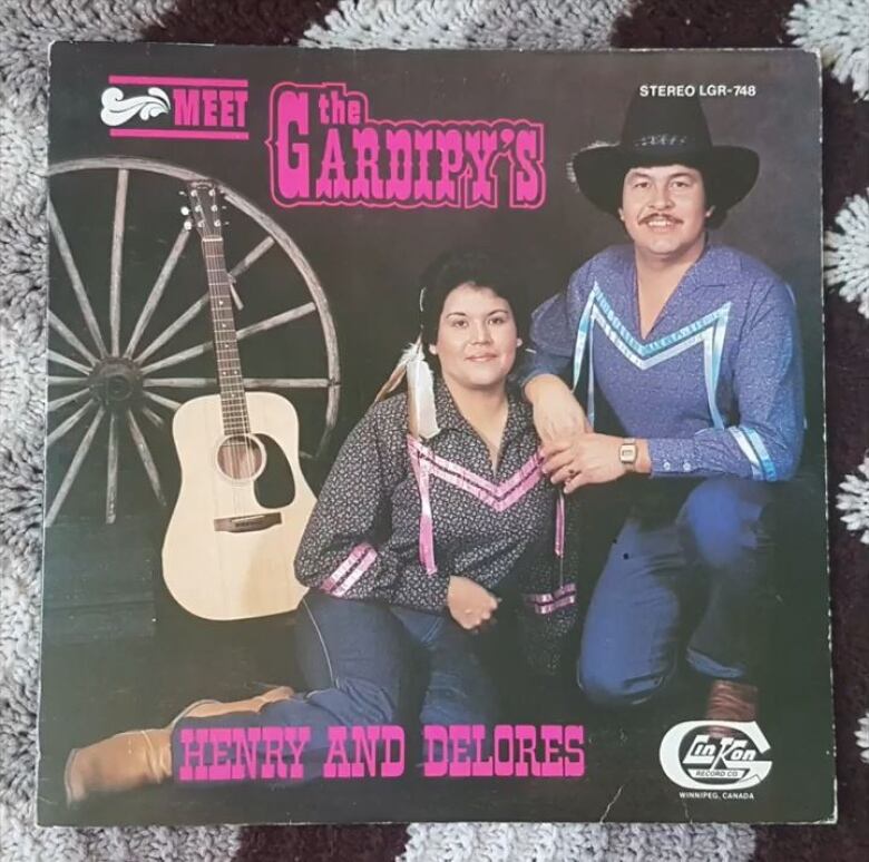 A wagon wheel and acoustic guitar sits behind a husband and wife posing on an album cover