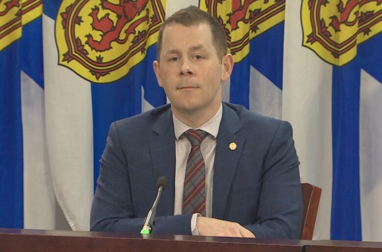 A man with a fauxhawk wearing a suit sits at a podium backed by Nova Scotia flags.