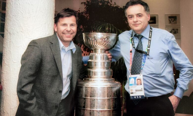 Two men stand with a big silver trophy, the Stanley Cup. One is smiling broadly in a sports jacket and buttoned shirt with an open collar, the other has a slight smile and a shirt and tie, with a large official badge hanging from his neck. 