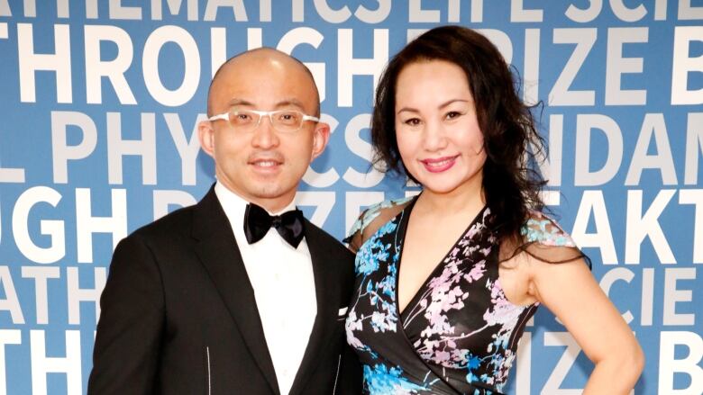 A man in a dark tuxedo and a woman in a dress are shown standing on a red carpet at an event.