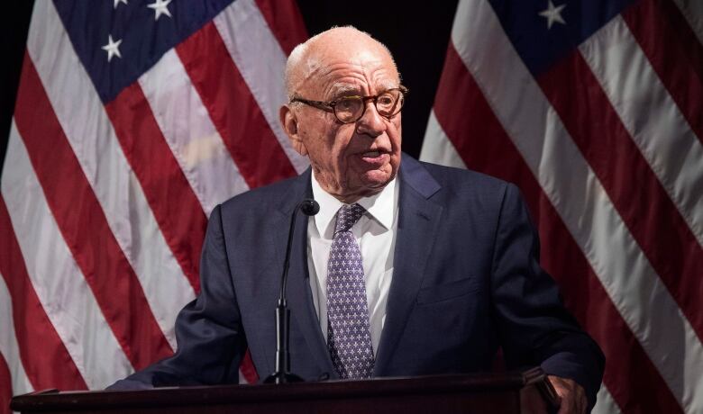 An older man is seen in a suit speaking at a podium in front of several American flags.