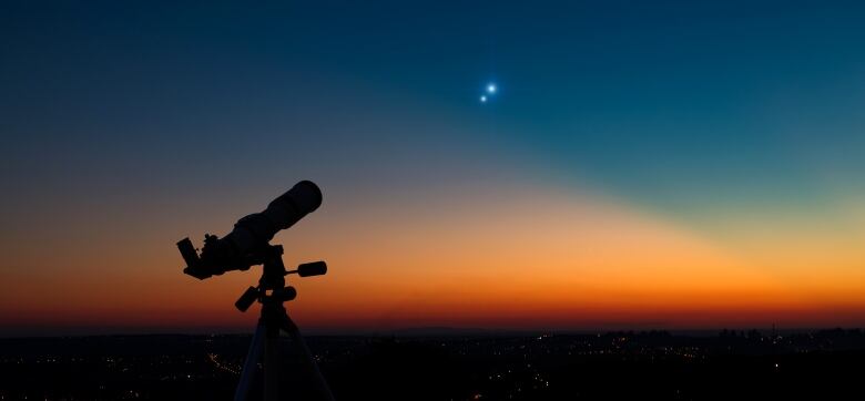 A telescope is silhouetted against an evening sky, with two bright star-like objects that are actually Jupiter and Saturn.