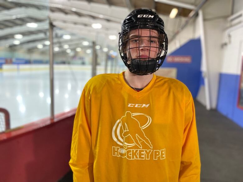 A hockey player wearing a yellow jersey and black helmet stands next to the ice at a rink