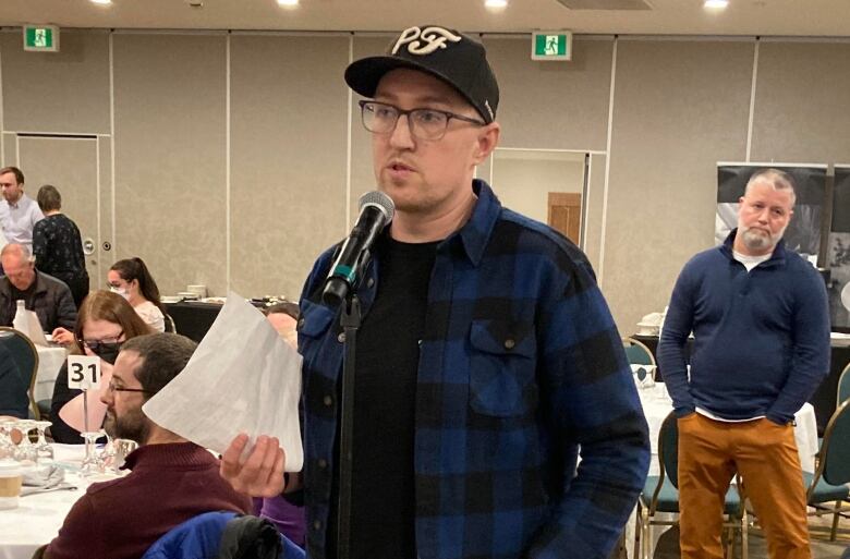 A man wearing a baseball hat speaking into a microphone and holding papers in his hand.