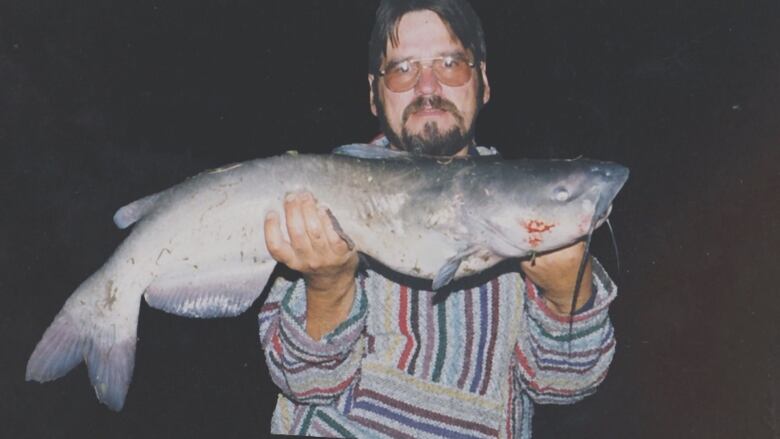 A vintage photo of a man hold a catfish up to the camera is shown.