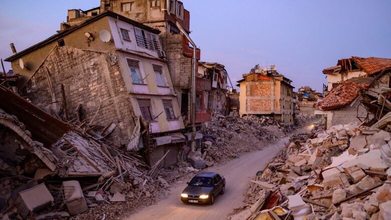 A car drives past collapsed buildings.