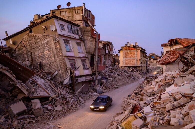 A car drives past collapsed buildings.