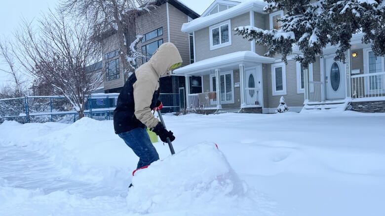 A man in a winter coat and hood shovels snow from a sidewalk.