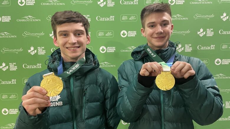 Two smiling young men with short hair and wearing dark green puffy coats hold out the orange-sized gold medals hanging from wide blue, green and white variegated, ribbons around their necks.