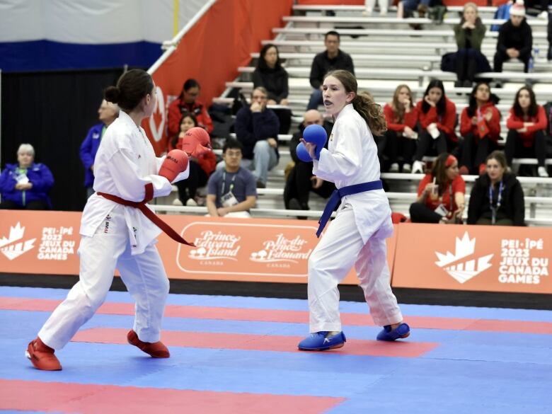 Two young women in white Gis face off against each other on a blue and red mat. One faces away from the camera wearing red shoes, gloves and belt. She has short, dark hair. One faces towards the camera wearing blue shoes, gloves and belt. She has long blonde hair in a pony tail. About 20 fans can be seen seated sporadically on several rows of bleachers in the background.