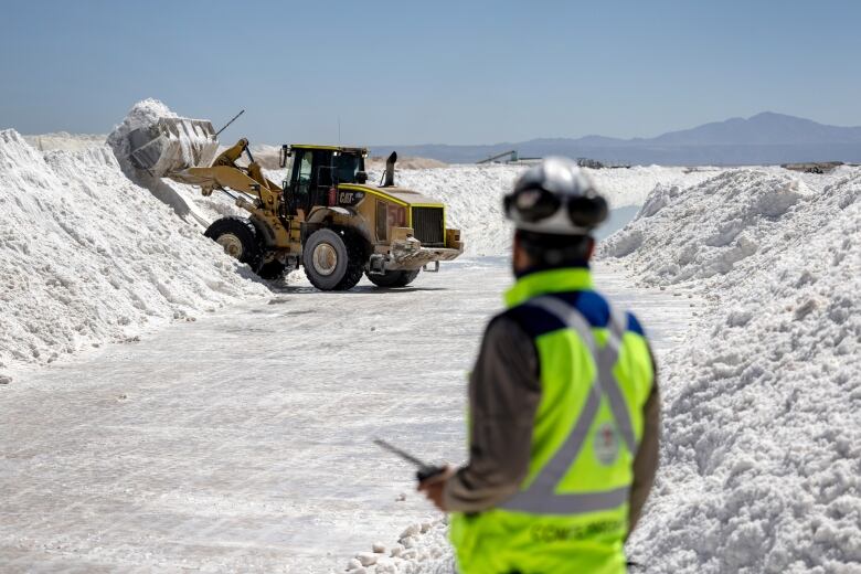 Labourer wearing flourescent safety vest stands in the foreground, looking at a dozer dumping lithium onto a piled row of the metal.