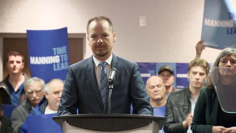 A man in a blue suit stands in front of a podium in front of people holding signs.