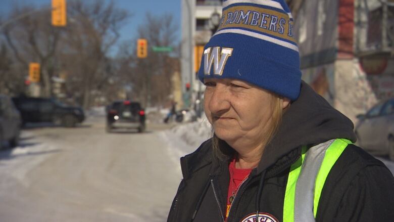 A woman in a Winnipeg Blue Bombers toque and a neon vest and black hoodie stands near a street.