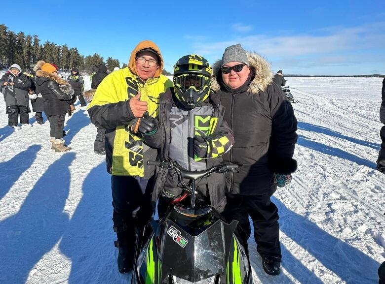 A mother and father stand next to their son on a snowmobile in a wide open expanse of a snow covered lake.