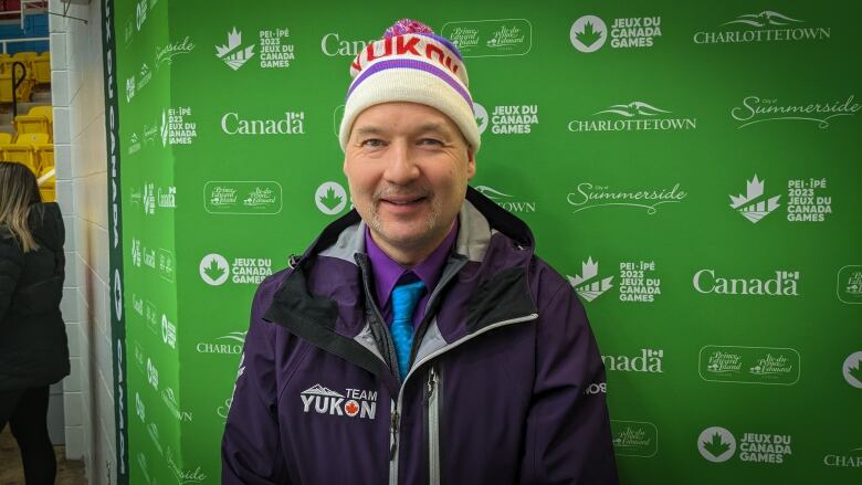 A man with a gray beard poses for a picture while wearing a purple coat and a white beanie with the word 