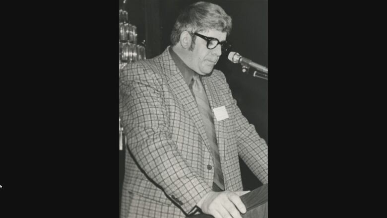 A black and white photo of a man with dark hair wearing a suit speaking at a podium