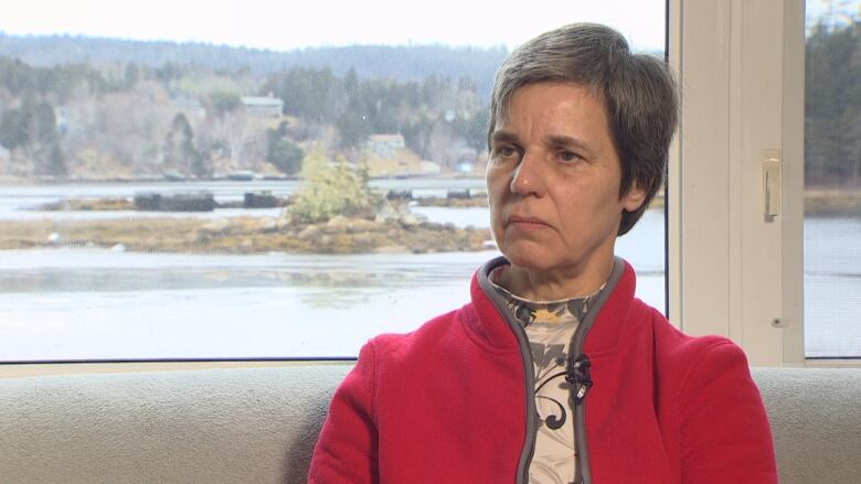 A woman in a red sweater sits in front of a window that looks out onto a cove.