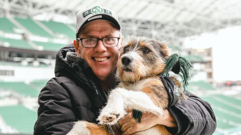 Man poses holding white, brown and black dog in the stands of football stadium