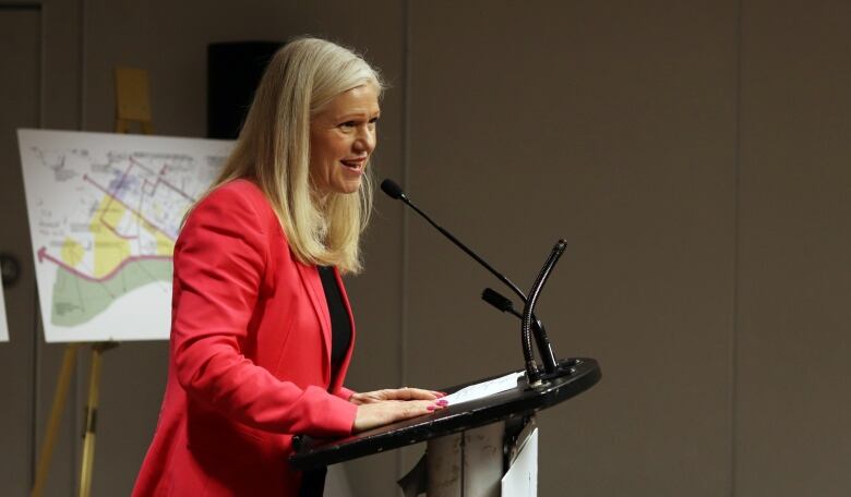 A woman with blond hair wearing a red blazer at a podium with a poster board showing downtown Moncton in the background.