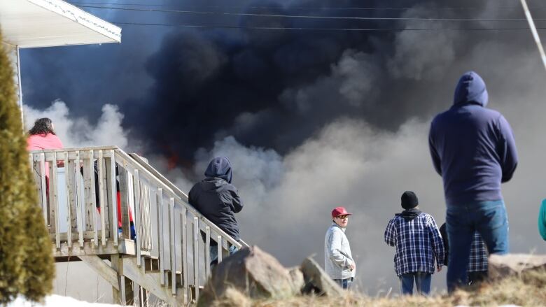 People stand on steps to a house and on sidewalk and watch as dark smoke billows from a nearby house fire.