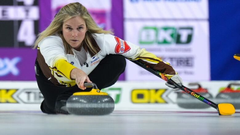 A curler holds her broom in her left hand as she slides across the ice readying to throw a yellow rock in her right hand.