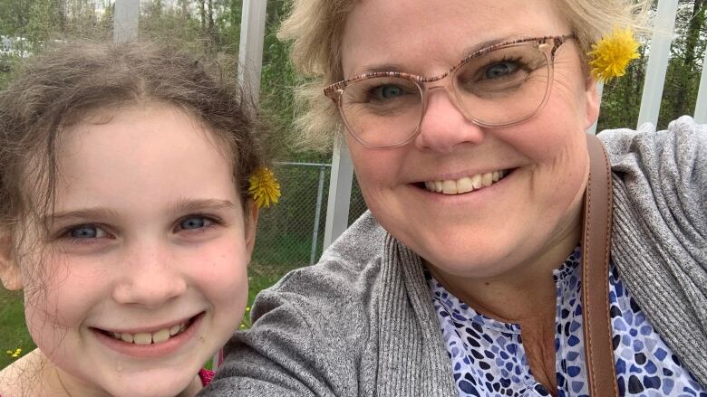 A woman in her 50s with short blonde hair and eye glasses and girl with brown hair in a pony tail look at the camera and smile. They each have a dandelion tucked into the hair behind one ear.