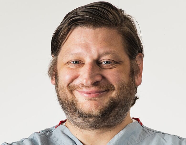 A man in his 40s with short brown hair parted on the side and a short beard and mustache smiles at the camera for a head and shoulders portrait wearing a pale blue surgical scrub shirt.