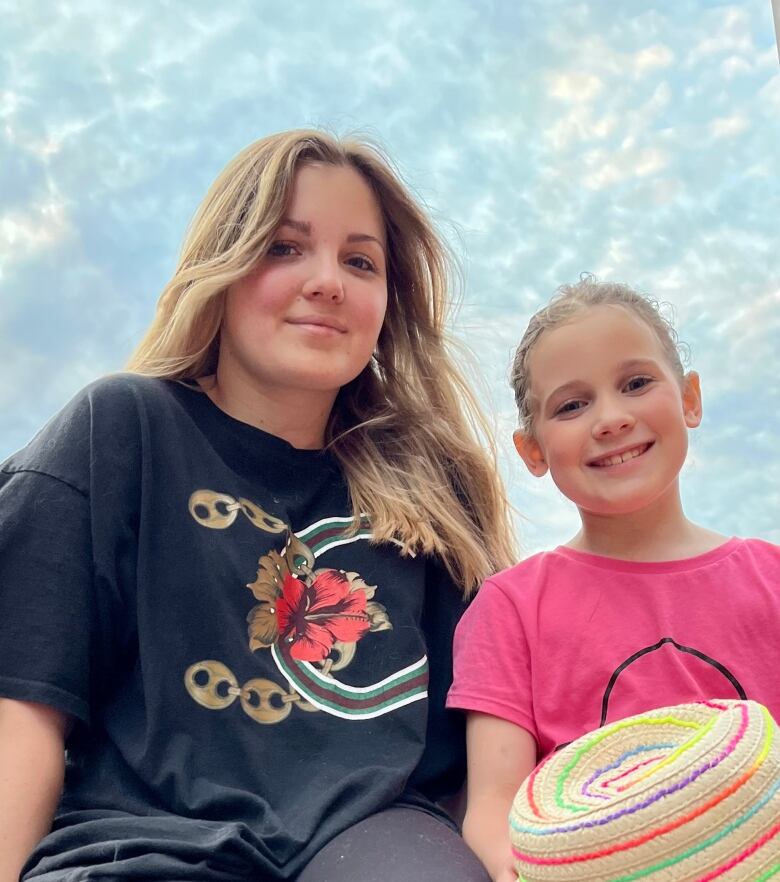 A teen with long light brown hair and wearing a black t-shirt and a pre-teen wearing her blonde hair parted in the middle and tied back and wearing a bubble gum pink t-shirt and holding a straw hat with Easer-coloured stripes on it smile at the camera at a downward angle against a pale blue sky with cirrocumulus clouds.