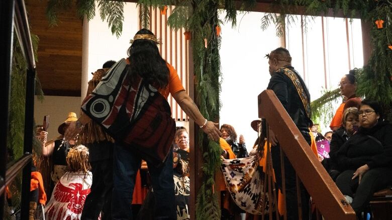 A group of people in First Nations regalia stand in a circle. Above them, cedar boughs hang, as well as small orange shirts cut out of paper. 