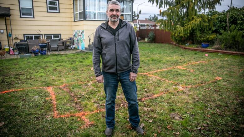 A man with a beard stands in his yard. Orange paint marks plans for a bungalow for his kids to move into.