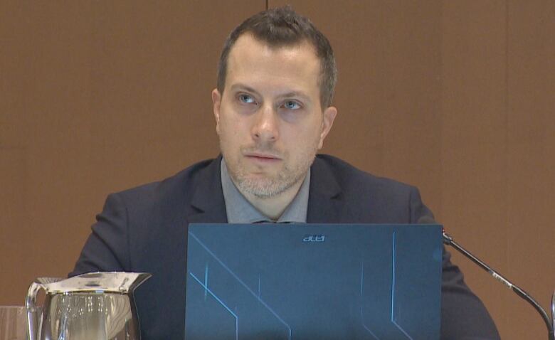 A man with short brown hair sitting in front of a laptop. There is a silver pitcher on either side of him and a disposable coffee cup on the table on the left.