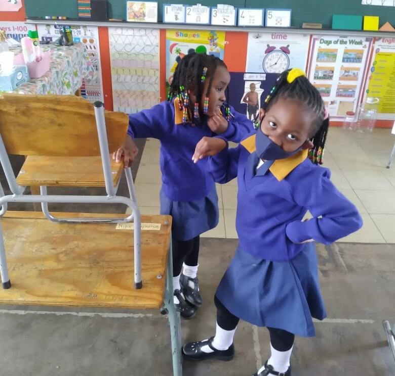 Two girls with braids and dressed in blue uniforms inside a classroom with colourful posters on the wall.