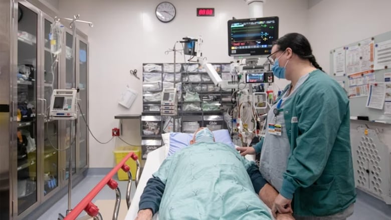 A masked female nurse holds the wrist of a male patient lying on a stretcher in an emergency room, surrounded by equipment and supplies.