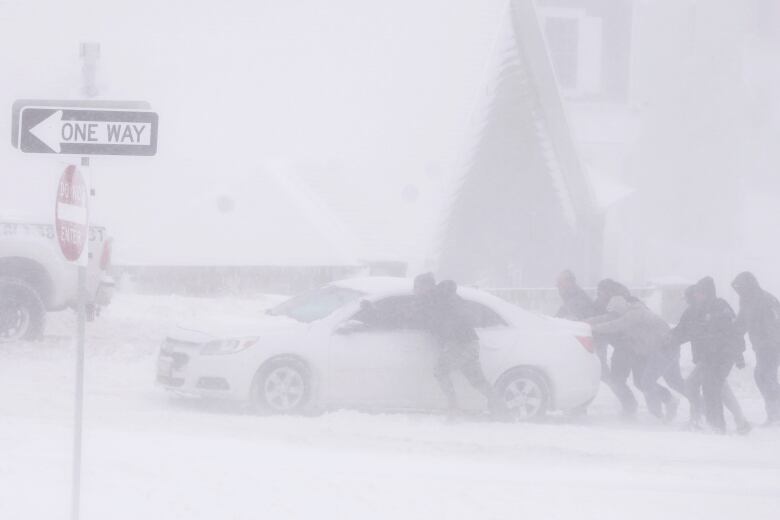 Several people are shown trying to push a car out of the snow, in blustery conditions.