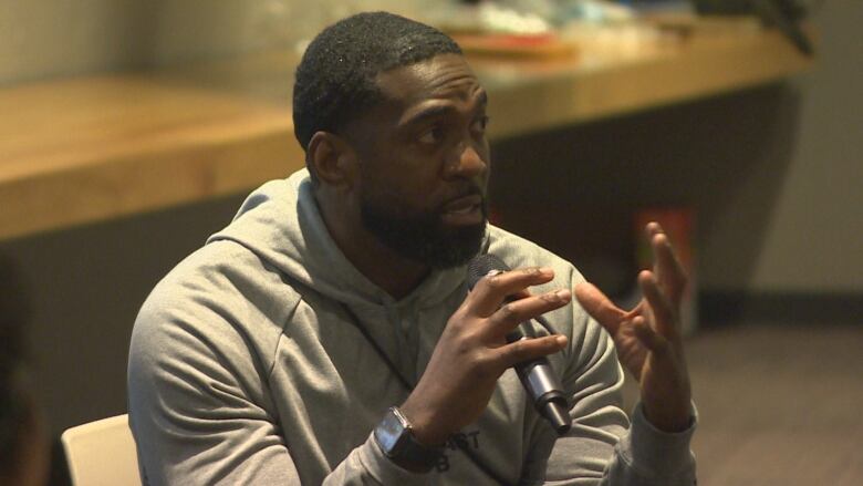 A man speaks into a microphone he's holding while sitting down.