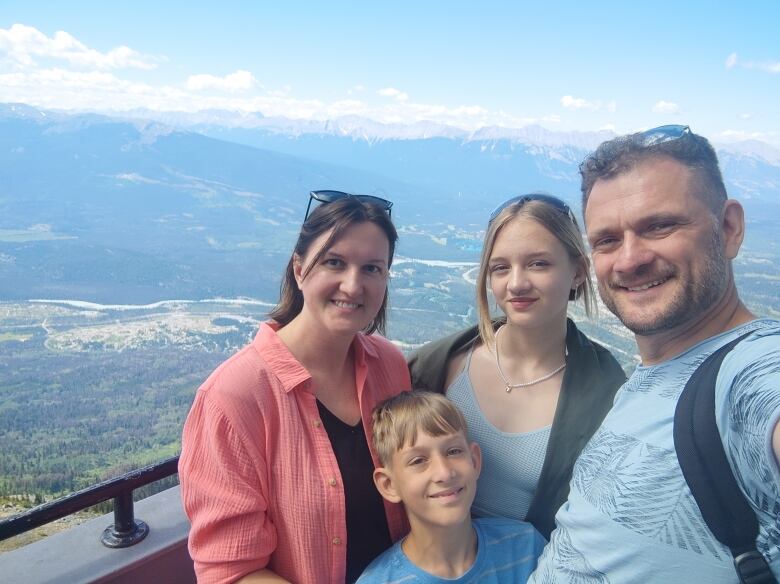 A family of four is taking a selfie on a lookout. The lookout oversees a valley and there are mountains in the background.