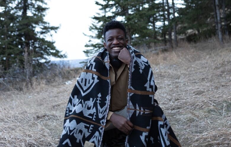 A man sits in a forest, smiling, with a blanket wrapped around him.