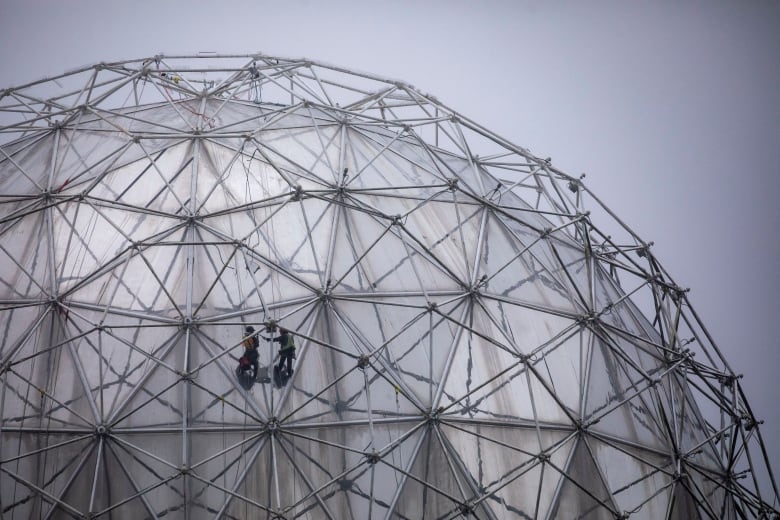 Suspended workers are pictured repairing lights on the outside of the silver Telus Science World dome on a cloudy day.