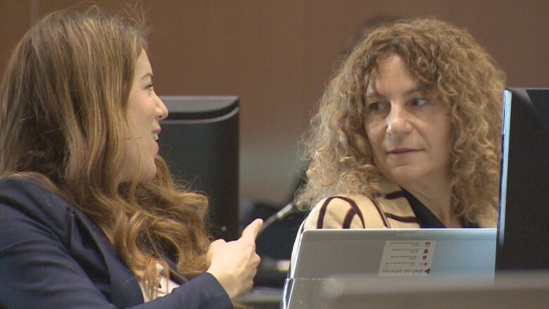 Two woman talk behind computers wearing business attire.