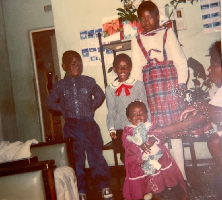 A smiling toddler holding a stuffed toy with her mother reaching toward her from the side poses in front of two older children, and a young woman. 