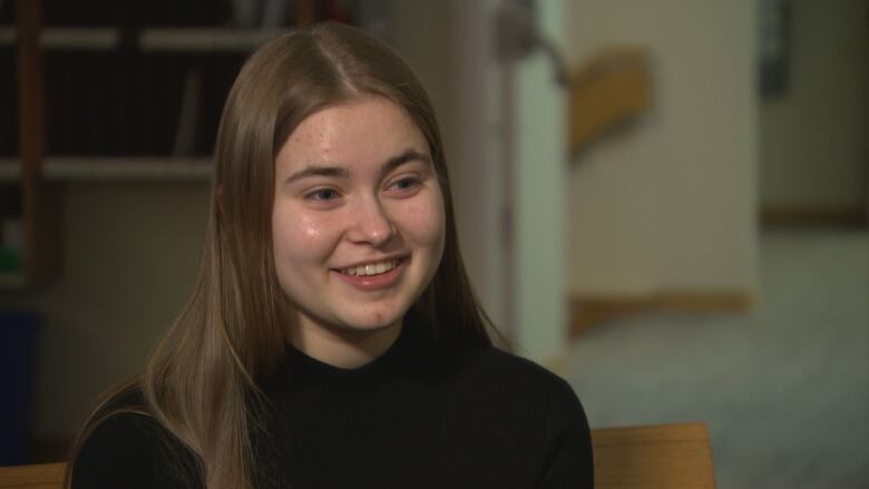 A teenage girl in a black turtleneck smiles and looks forward. 